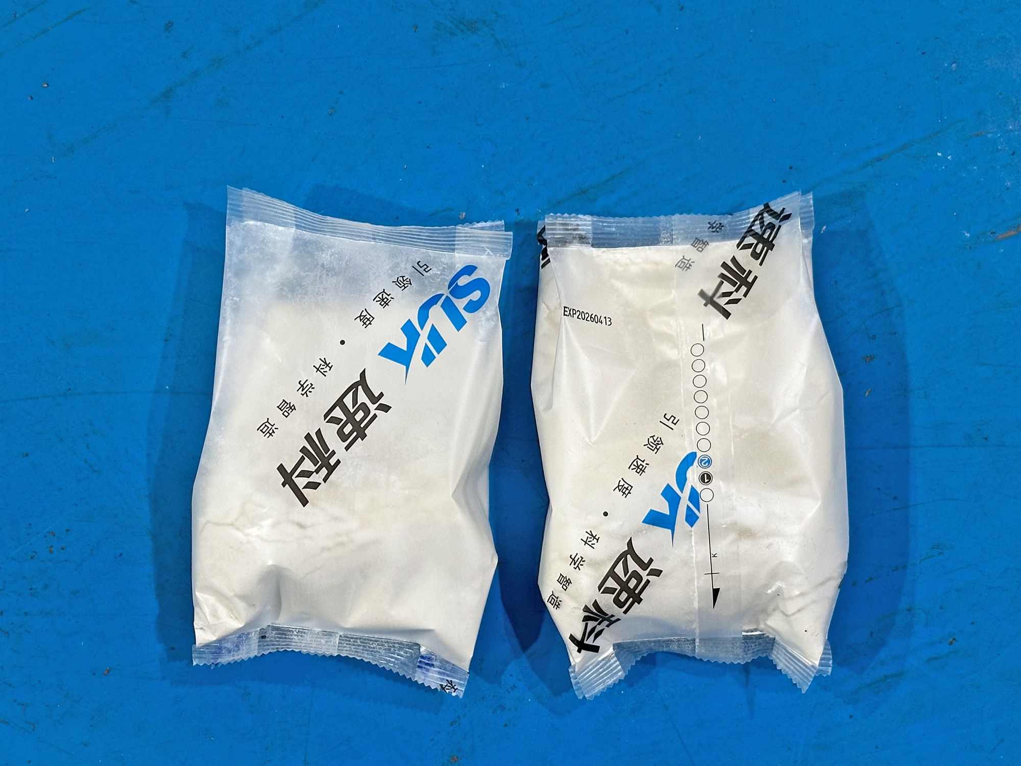 Two sealed white powder pouches showing the seal area and printed panel on a blue factory floor